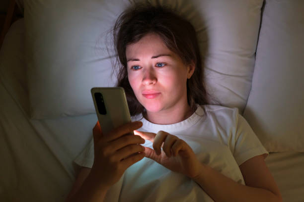 Portrait of a young woman who lies on the bed at night and types messages on a cell phone. stock photo