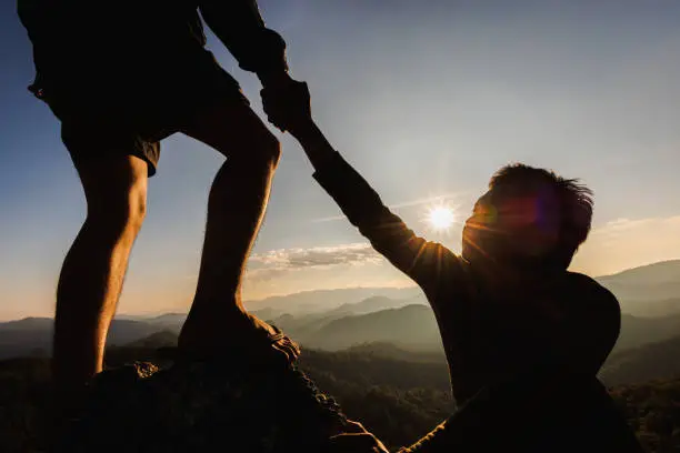 Photo of Silhouette of hiker helping each other hike up a mountain at sunset. People helping and, team work concept.