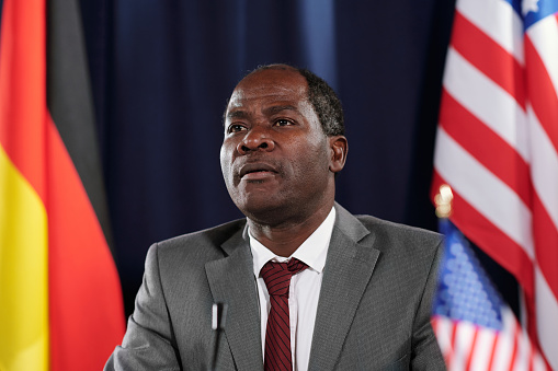 African American male delegate in elegant suit sitting in front of camera against flags of germany and USA taking part in political summit