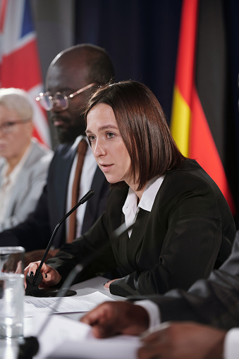 Young female politician in formalwear speaking in microphone while sitting among intercultural foreign colleagues at conference or summit