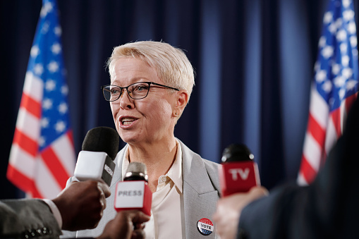 Blond mature female deputy or American delegate speaking in microphones held by hands of journalists at press conference