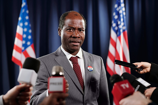 Mature African American deputy in formalwear speaking in microphones of various television channels during press conference or interview
