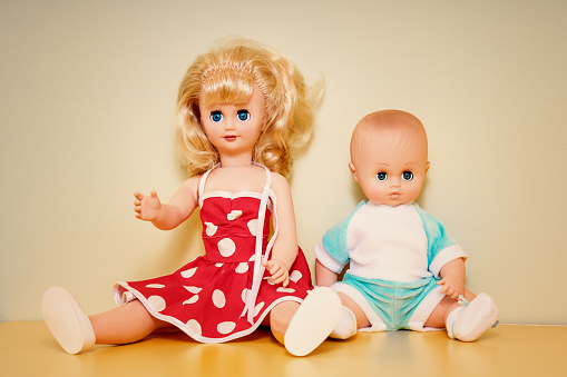 Couple of old retro plastic dolls seated on wooden table against concrete wall. Antique vintage children playthings concept.