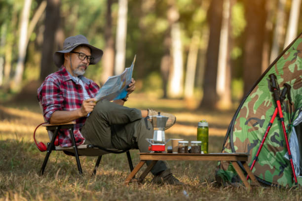 istock Happy middle-aged Asian man drinking coffee holding a map in a campsite next to a tent. Alone in the forest with sunlight on the hill 1471817849