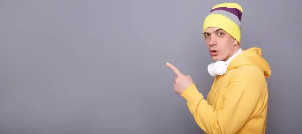 Side view portrait of surprised astonished man in beanie hat and yellow casual hoodie, pointing away at copy space, advertisement area, freespace for promotion, standing isolated over gray background.