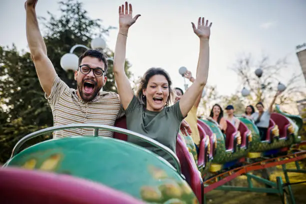 Happy couple having fun while riding on rollercoaster with raised arms at amusement park.