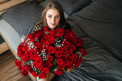 A beautiful girl is sitting on a bed in the bedroom with a huge bouquet of scarlet roses. Flower delivery to women on holidays and memorable dates.
