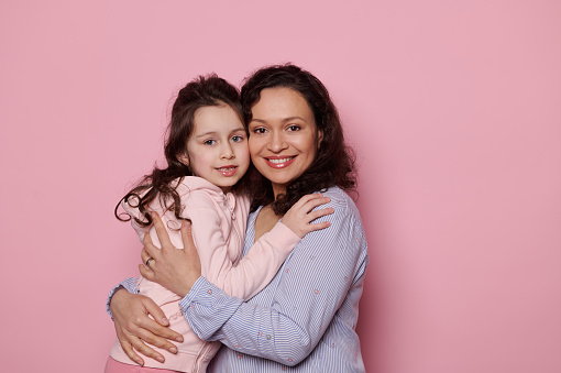 Loving mother hugging her little daughter, cutely smiling looking at camera, standing together on isolated pink background. Copy space. Happy family relationships. Mother's and Children's Day concept