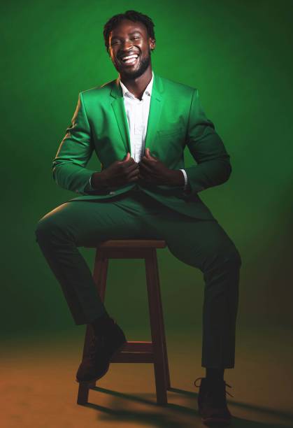 Fashion, trendy and black man model in a suit on chair in the studio with elegant, fancy and stylish outfit. Happy, smile and portrait of African male with formal clothes isolated by green background Fashion, trendy and black man model in a suit on chair in the studio with elegant, fancy and stylish outfit. Happy, smile and portrait of African male with formal clothes isolated by green background dinner jacket stock pictures, royalty-free photos & images