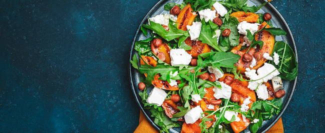 Summer salad with grilled peach with soft cheese, hazelnuts and arugula on blue table background, top view, copy space