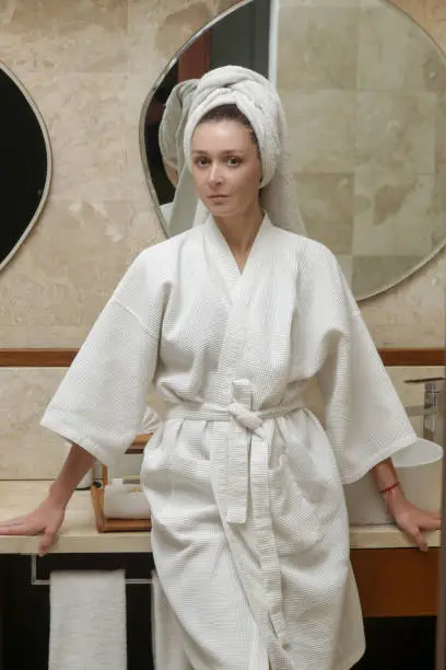 Portrait of young woman in white cotton bathrobe and hair wrapped in towel in the bathroom