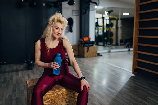 Portrait of a carefree middle-aged woman sitting on a box at the gym, holding a bottle in her hands