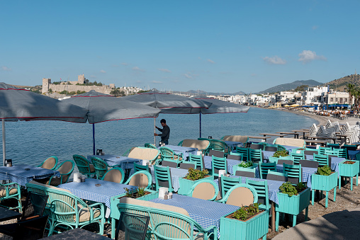 Bodrum, Mugla, Turkey. April 21st 2022\nA waiter prepares tables on the beach side cafe in Bodrum harbour with Bodrum Castle in the background, the Aegean coast of south west Turkey.