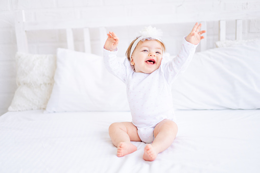 funny happy baby girl sitting on the bed in white clothes and holding her hands up dancing or stretching after sleeping in the morning, a small child on a cotton bed at home smiling