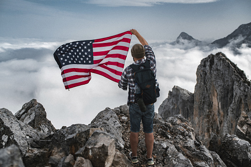 Proud man raising the flag of the United States of America standing on the top of the mountain above beautiful clouds. Successful climb to the top of the mountain.