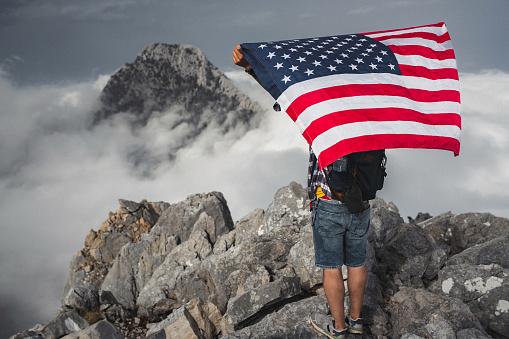Proud man raising the flag of the United States of America standing on the top of the mountain above beautiful clouds. Successful climb to the top of the mountain.