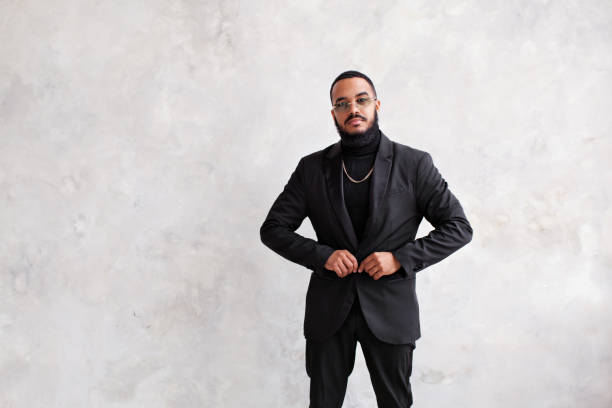 istock Handsome young arabian man with beard buttoning up jacket, wearing black suit and turtleneck against concrete wall. Total black male look 1471842545