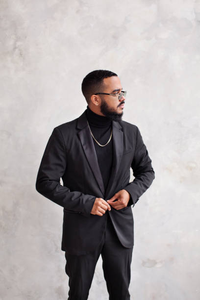 istock Handsome bearded man buttoning up black jacket, wearing stylish suit, glasses and turtleneck against concrete wall. Total black fashion male clothes 1471842547