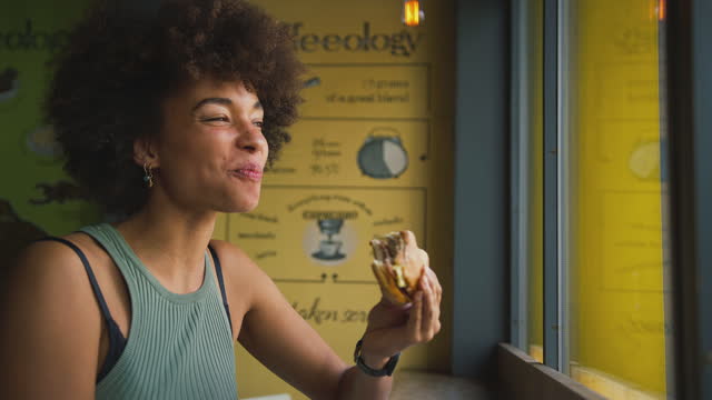 Female Customer In Coffee Shop Window Eating Bagel Sandwich For Lunch
