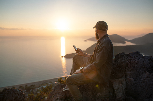 Adult man in modern clothing sitting on the top of the mountain with smart phone and watching sunset at sea from the top.