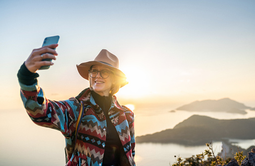 Happy cheerful woman in modern outfit taking selfies against beautiful beach at sunset. Oludeniz Beach, Turkey