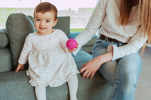 Toddler baby sitting on the side of couch with her mother. A child girl is standing on the sofa with a toy on her hand. Kid age one year.