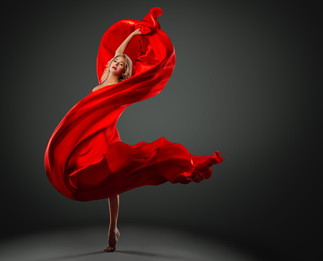Ballerina dancing with Red Silk Fabric flying on Wind. Modern Ballet Dancer jumping over Dark Studio background. Fashion Woman in Red Dress wrapped in waving Scarf