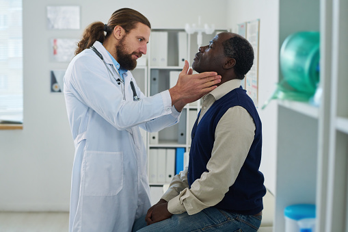 Young endocrinologist checking thyroid of mature African American male patient in casualwear sitting by wall in front of him in hospital