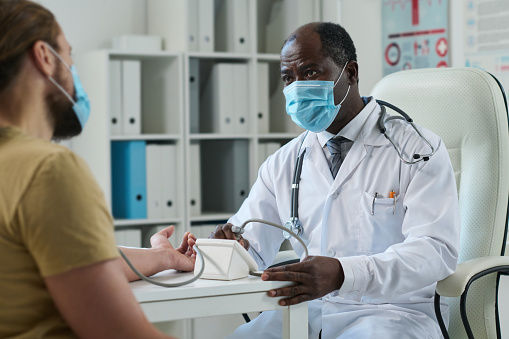 Mature male doctor in protective masks looking at young patient while checking up his blood pressure with tonometer in clinics