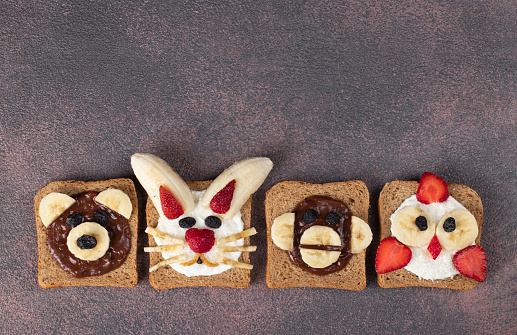 Four sweet toasts for kids in shape of chick, bear, rabbit and monkey, with strawberries, banana, cream cheese, chocolate and coconut flakes on brown background, Top view