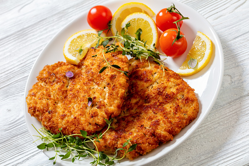 fried breaded chicken breast cutlets on white plate with lemon, fresh thyme and ripe tomatoes on textured wood table, close-up
