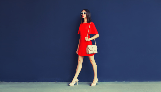 Beautiful young full-length woman in red dress with handbag and heels on blue background