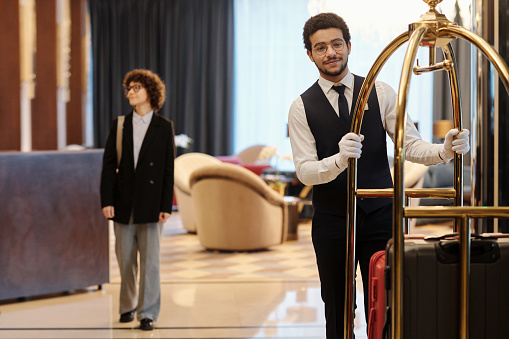 Young bellboy in suit pushing cart with many suitcases of hotel guests while moving along lounge with female traveler on background