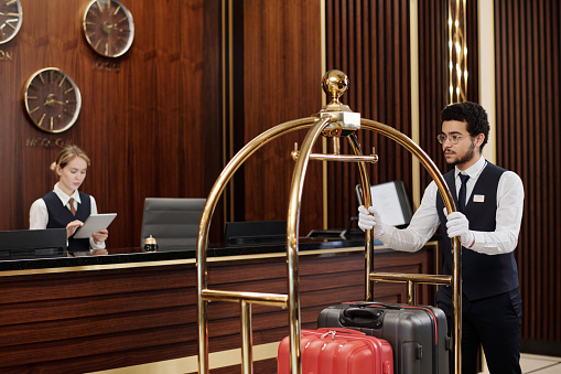 Young bellboy in uniform pushing cart with suitcases while walking along reception counter with female manager using digital tablet