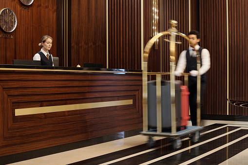 Blurry motion of bellboy pushing cart with clients luggage while walking along reception counter with young female manager in uniform