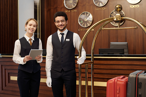 Happy young elegant receptionist and bellboy standing in lounge of luxurious hotel and looking at camera while waiting for new guests