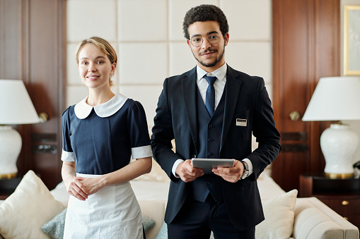 Young successful staff of luxurious five star hotel standing against couch with cushions and two lamps in room prepared for new guests