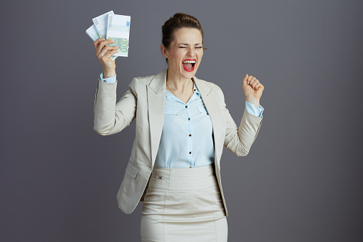 happy elegant small business owner woman in a light business suit with euros money packs against gray background with raised arms rejoicing.