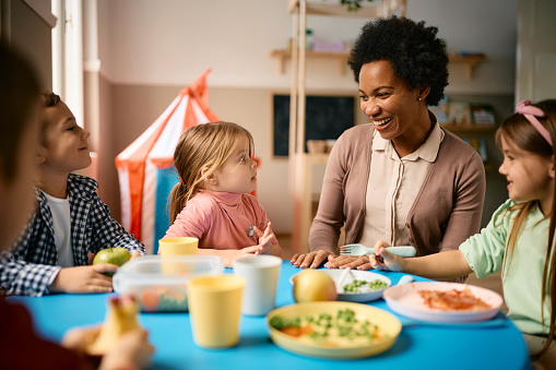 Group of happy kids communicating with their African American teacher while eating lunch at preschool.