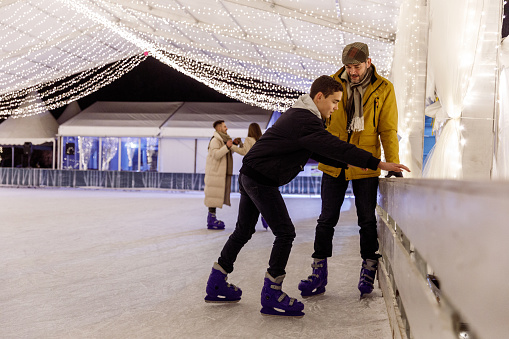 Candid shot of teenage boy showing his father and teaching him how to ice skate on their outing at the ice skating rink. Couple holding hands and ice skating in the background.