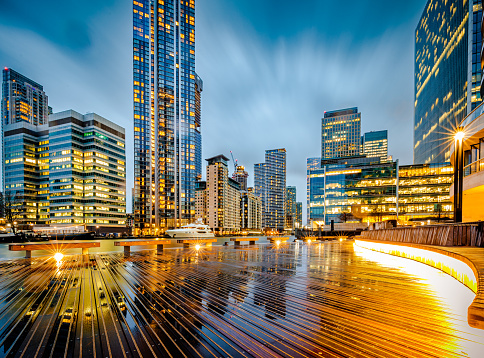 The skyscrapers and high rise apartment buildings of London's financial district at Canary Wharf on the Isle of Dogs. The modern cityscape is illuminated in the blue hour just after sunset with the buildings reflected in the wet wooden decking in the foreground.