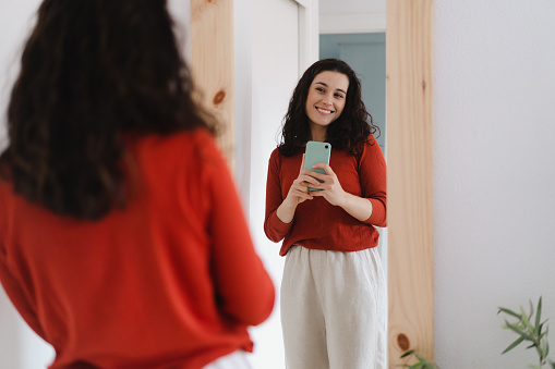Young beautiful woman in red sweater taking a selfie with the smartphone of her reflection in a mirror