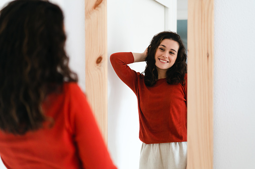 Young beautiful self confident woman in red sweater looking in the mirror proud of herself