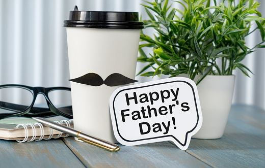 Happy fathers day concept. coffee cup with paper, heart tag with Happy father's day text and notebook, glasses on wooden table background.