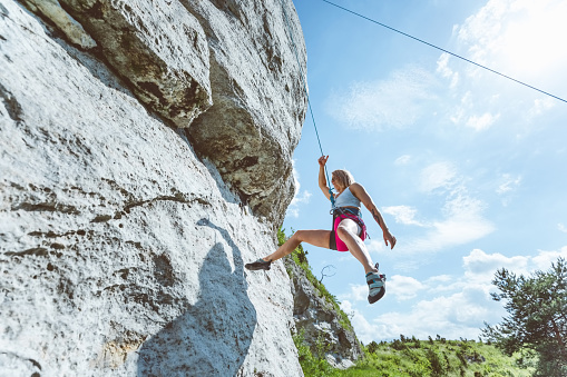 Wide angle view of young woman climbing on cliff rock with cloudy sky in the background, hanging on rope.