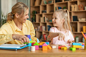 Positive kids psychology. Professional female therapist with notepad watching little girl playing with colorful blocks