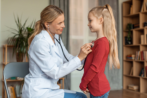 Cheerful female pediatrician listening girl's heartbeat with stethoscope during checkup at home, pediatrist doctor checking child's heart and lungs, side view