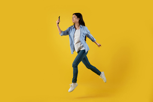 Glad millennial asian female in casual froze in air jumping and look at smartphone, isolated on yellow background, full length, profile. Selfie for blog and social networks, modern technology and app