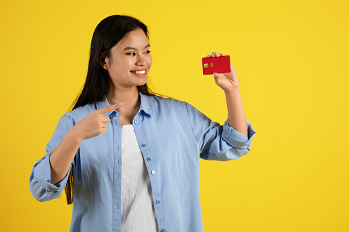 Cheerful happy young japanese female pointing finger at credit card, recommends save money, isolated on yellow background, studio. Shopaholic and shopping, sale, finance and payment of purchases