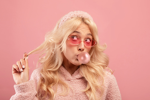 Pretty blonde lady blowing bubble from chewing gum, looking aside and twisting lock of hair on her finger over pink studio background. Stylish woman in hoodie and glasses having sweet tooth